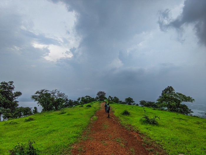 Two hikers on a red earth trail along a green ridge under heavy monsoon cloud