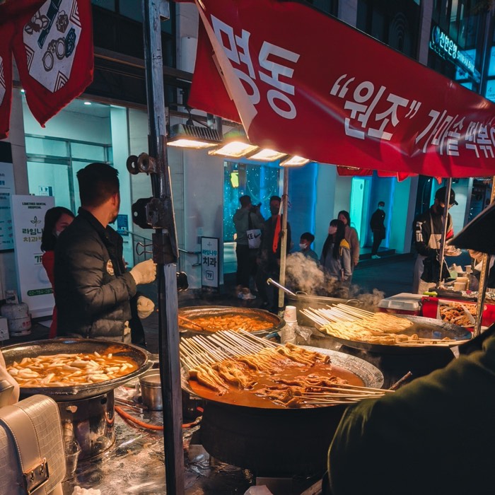 Steam rising from pans of tteokbokki and fish cakes under a red Myeongdong awning