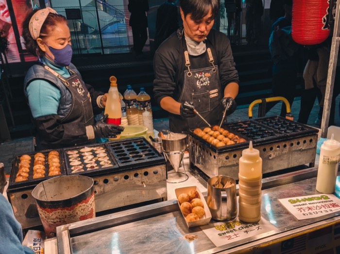 Two vendors turning takoyaki on hot griddle plates at a night stall