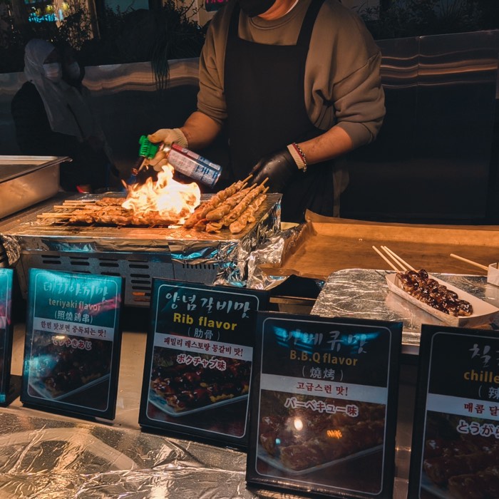 Skewers being flame-torched at a street stall, flavour signs propped along the counter