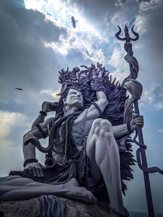 Towering stone statue of Shiva holding a trident, shot from below against dramatic clouds